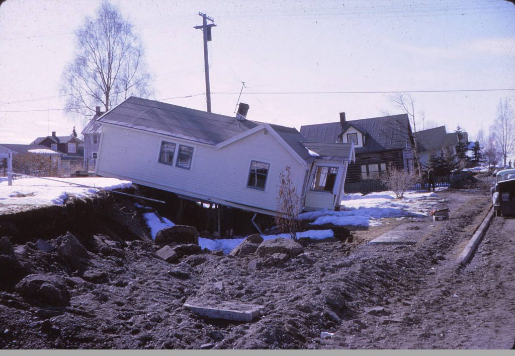 A view of a house damaged by ground dropping near downtown Anchorage after the 1964 earthquake. The fronts of houses further down the street seem to be tilted upward.Bowles family papers, Archives and Special Collections, Consortium Library, University of Alaska Anchorage.